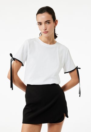 Weißes Baumwoll-T-Shirt mit kurzen Ärmeln, das schwarze Bänderakzente an den Ärmeln aufweist, kombiniert mit einem figurbetonten schwarzen Minirock.
