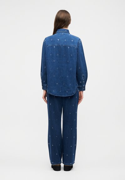 Femme vue de dos portant une chemise en jean bleue assortie et un pantalon avec de petits motifs blancs brodés, debout sur un fond blanc.