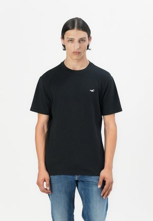 Black cotton t-shirt with short sleeves and a crew neck. Features a small white logo on the left chest. Paired with blue jeans.