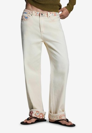 Personne portant un jean droit couleur crème avec des poignets et une ceinture à motifs, associé à des tongs noires et un pull vert olive.