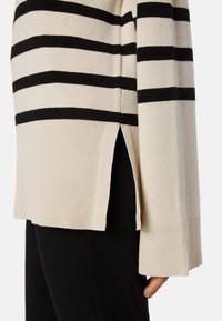 Gestreifter Pullover in Creme und Schwarz, mit ribbeltem Saum, seitlichen Schlitzen und weichem Strickmaterial. Detaillierte Ansicht des Stoffes und des Schnitts.