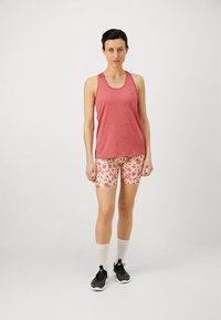 Punainen hihaton toppi väljän mallin kanssa, yhdistetty beigeihin kukkakuosisiin shortseihin. Mallilla mustat urheilukengät ja valkoiset sukat.