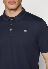 Granatowa koszulka polo z teksturowanej tkaniny, trzy guziki i małe białe logo "CK" na lewej piersi. Klasyczny krój kołnierza.