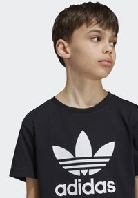 T-shirt en coton noir avec un col rond et des manches courtes, comportant un grand logo trefoil blanc d'Adidas sur le devant.