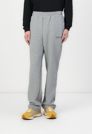 Pantaloni de trening gri cu talie elastică, având buzunare laterale și un logo imprimat pe partea stângă. Purtați cu adidași galbeni și gri.