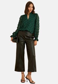 Zelená, průhledná blůza s texturovanými kruhovými akcenty, spárovaná s černými širokými kalhotami z umělé kůže s vysokým pasem a černými sandály na podpatku.