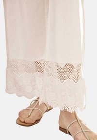 Pantalones blancos y ligeros con un borde de encaje en forma de ondas que presenta bordados florales y patrones de malla abierta en el dobladillo. Sandalias con tiras finas.