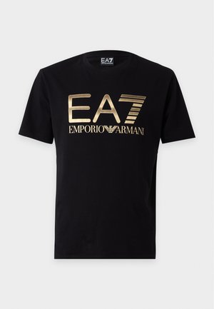 Mężczyzna w jasnobrązowej kurtce zapinanej na zamek błyskawiczny, oliwkowo zielonych dresach z białym logo EA7 Emporio Armani i czarnych trampkach, stoi z rękami w kieszeniach.