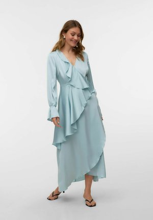 Frau trägt ein hellblaues, langärmeliges Wickelkleid mit Rüschen und schwarze Sandaletten mit Absatz, steht vor einem schlichten weißen Hintergrund.