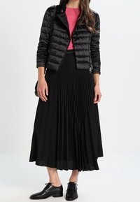 Femme portant une doudoune noire, un haut rose, une jupe midi plissée noire, des chaussures en cuir noires et portant un sac à bandoulière noir.