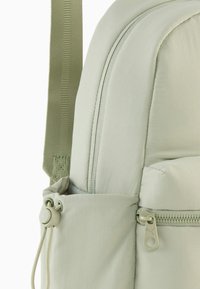 Sac à dos gris clair en tissu doux avec une texture lisse, doté d'une poche zippée et de bretelles vertes ajustables.