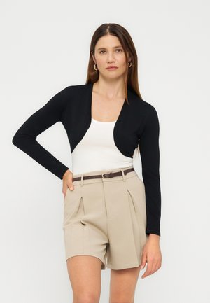 Femme portant un shrug noir court à manches longues sur un haut blanc avec un short beige taille haute plissé et une ceinture marron, posant avec la main sur la hanche.