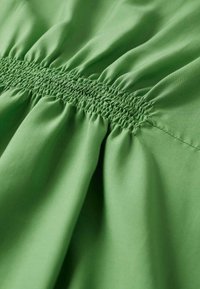 Tessuto verde con una texture arricciata, caratterizzato da una sezione smock. Il materiale è liscio e leggero, creando morbide pieghe fluide.