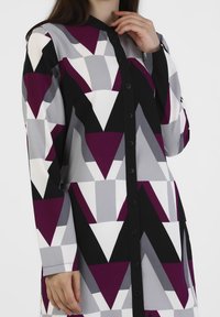 Robe à manches longues présentant un motif géométrique dans des tons de violet, noir et gris. Confectionnée en tissu texturé avec des fermetures à boutons.