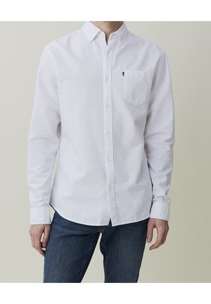 Homme portant une chemise blanche à manches longues avec poche poitrine et un jean bleu foncé sur un fond uni.