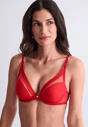 Femme aux longs cheveux foncés portant un soutien-gorge rouge avec bordure en dentelle et détails en mesh, regardant légèrement de côté sur un fond uni.