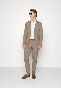 Light brown suit with a fitted blazer and slim trousers, paired with a white patterned shirt and brown shoes. Accessories include dark sunglasses.