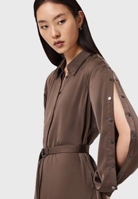 Vestito camicia in satin marrone con scollatura a collo, maniche lunghe con dettagli a pressione e vita con cintura. Tessuto liscio e vestibilità sagomata.