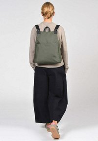 Mochila verde oliva con diseño cuadrado, fabricada en tela, con correas negras y un bolsillo frontal. Llevada con pantalones holgados de color carbón.