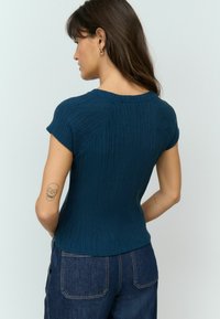 Dunkeltürkises Strick-T-Shirt mit kurzen Ärmeln, strukturiertem Rippmuster, rundem Ausschnitt und enganliegendem Design, kombiniert mit blauen Jeans.