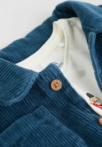 Chaqueta de tela de pana azul con una textura suave y acanalada, que presenta un botón de madera y una capa interior blanca con un diseño bordado.