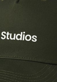Donkergroene pet van textuurstof met het woord "Studios" in vetgedrukte witte letters. Close-up toont de stiksel- en stofdetails.