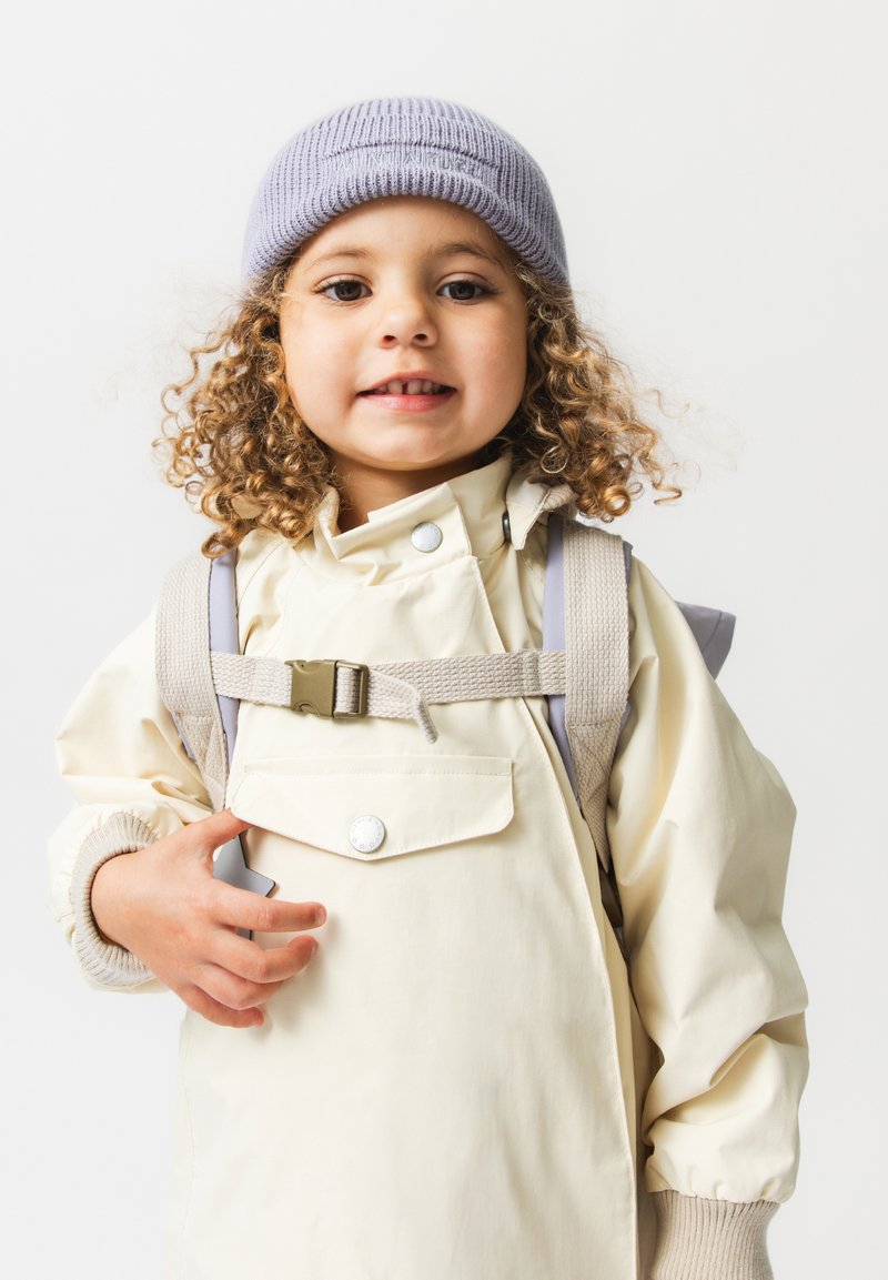 Μικρό παιδί με σγουρά μαλλιά που φοράει γκρίζο σκούφο και κρεμ μπουφάν, με σακίδιο στην πλάτη, που χαμογελά στην κάμερα.