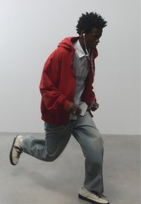 Fiatal férfi piros kapucnis pulóverben és farmerben, vezetékkel ellátott fülhallgatóval fut bent, egyszínű szürke háttér előtt.