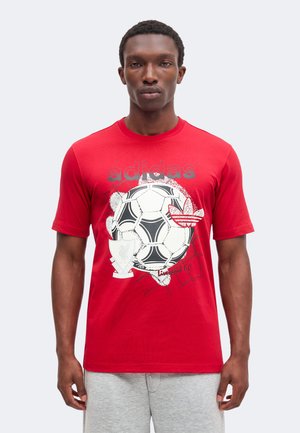 Άνδρας που φοράει κόκκινο μπλουζάκι Adidas με μεγάλη ασπρόμαυρη ποδοσφαιρική μπάλα, λογότυπο Adidas και σήμα της Liverpool F.C., συνδυασμένο με γκρι σορτς.