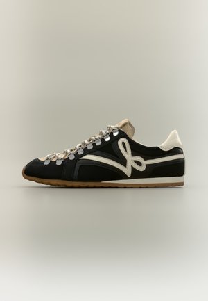 Baskets basses noires avec lacets crème et grand logo crème sur le côté, présentées de profil sur un fond uni.