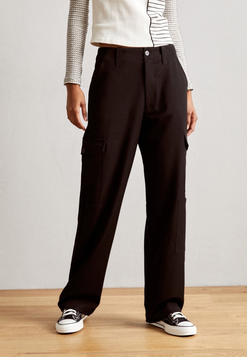 Personne portant un pantalon cargo noir ample avec des poches latérales, des baskets noires et blanches, et un haut blanc à manches longues avec des rayures noires.