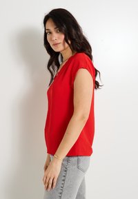 Haut rouge à manches courtes avec un décolleté en V, fabriqué à partir d'un tissu texturé, associé à un jean gris clair. La coupe est décontractée et légèrement ample.