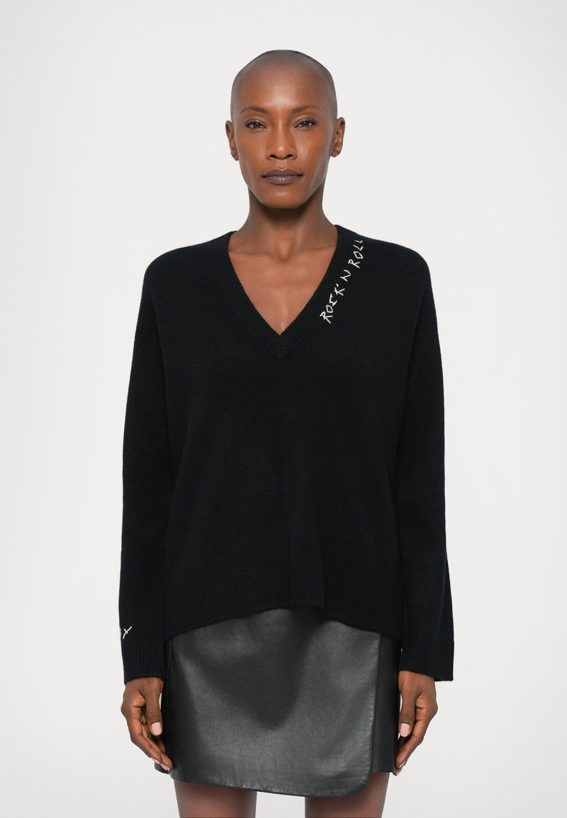 Schwarzer V-Ausschnitt-Pullover mit langen Ärmeln, versehen mit weißer "Rock 'N' Roll" Bestickung auf der Schulter. Kombiniert mit einem schwarzen Lederrock.