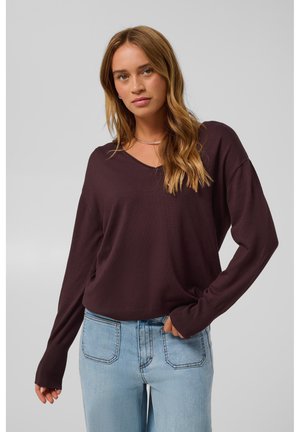 Jeune femme aux longs cheveux ondulés portant un pull violet foncé à manches longues et un jean bleu clair sur un fond uni.