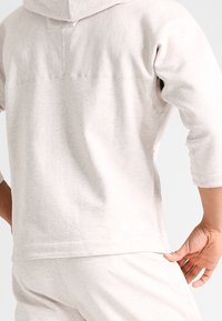 Sudadera beige clara de tela suave, con un bolsillo tipo canguro, puños acanalados y un dobladillo redondeado. La vista posterior muestra un diseño simple sin gráficos.