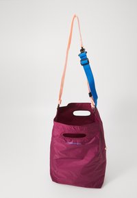 Složiva maroon torba s izrezanim ručkama, breskvasta naramenica i podesiva plava traka vise protiv jednostavne pozadine.
