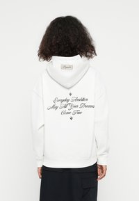 Haut à capuche blanc avec un grand texte brodé au dos en noir script. Comprend une capuche avec cordon de serrage et des poignets côtelés. Étiquette au col.