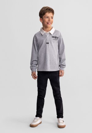 Lächelnder Junge steht, trägt ein graues Langarm-Poloshirt mit dem Schriftzug "UNLIMITED", schwarze Hose und weiße Sneakers vor hellem, einfarbigem Hintergrund.