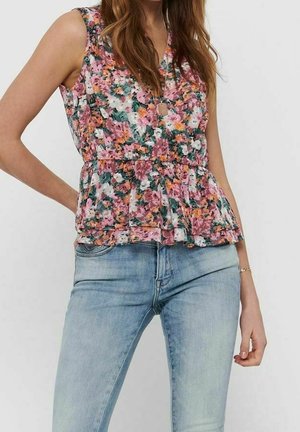 Femme portant un haut sans manches rose à motif floral avec des volants, associé à un jean en denim bleu clair, debout avec une main sur la hanche.