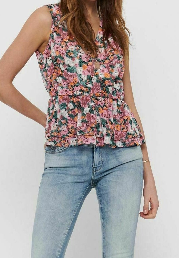 Femme portant un haut sans manches rose à motif floral avec des volants, associé à un jean en denim bleu clair, debout avec une main sur la hanche.