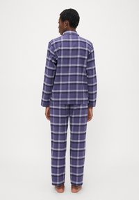 Persona de pie descalza, de espaldas, vistiendo un conjunto a juego de camisa de manga larga de cuadros morados y pantalones, contra un fondo liso.