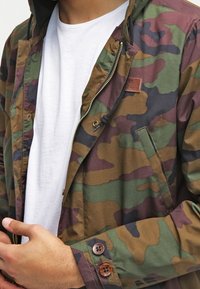 Muž v zeleno-hnědé maskáčové bundě přetažené přes bílé tričko, který si upravuje manžetu bundy levou rukou.