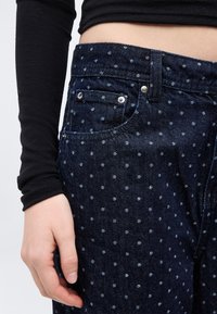 Κοντινό πλάνο ενός ατόμου που φορά σκουρό μπλε τζιν με μικρές ανοιχτό μπλε βούλες και μαύρο μακρυμάνικο τοπ, το χέρι χαλαρό δίπλα στο μηρό.