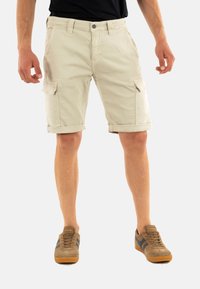 Pantaloni cargo khaki con orli arrotolati, tasche frontali e laterali, realizzati in tessuto di cotone, abbinati a sneaker marroni con strisce blu.