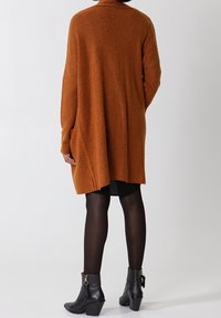 Pull orange surdimensionné avec une texture douce, dos de longueur moyenne et épaules tombantes. Porté sur une robe noire et des collants avec des bottines.