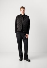 Black quilted vest with a zip closure and stand collar, layered over a black shirt, paired with loose black pants and black shoes.