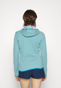 Persona che indossa una felpa con cappuccio di colore turchese e motivo, con spalline rosa di una canotta visibili, e pantaloni corti blu navy, vista da dietro su uno sfondo bianco.
