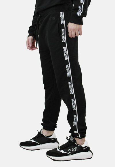 Pantaloni della tuta neri con logo Moschino bianco lungo i lati, dotati di un elastico in vita e polsini, abbinati a sneakers bianche e nere.