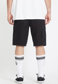 Schwarze Cargo-Shorts mit Seitentaschen, aus leichtem Stoff. Getragen mit weißen Kniestrümpfen mit schwarzen Streifen und schwarzen Schuhen.