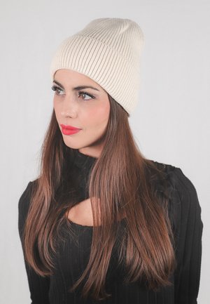 Berretto a coste crema, texture morbida, design aderente. Il modello indossa un maglione nero con dettagli a taglio. Capelli lunghi castani scendono oltre le spalle.